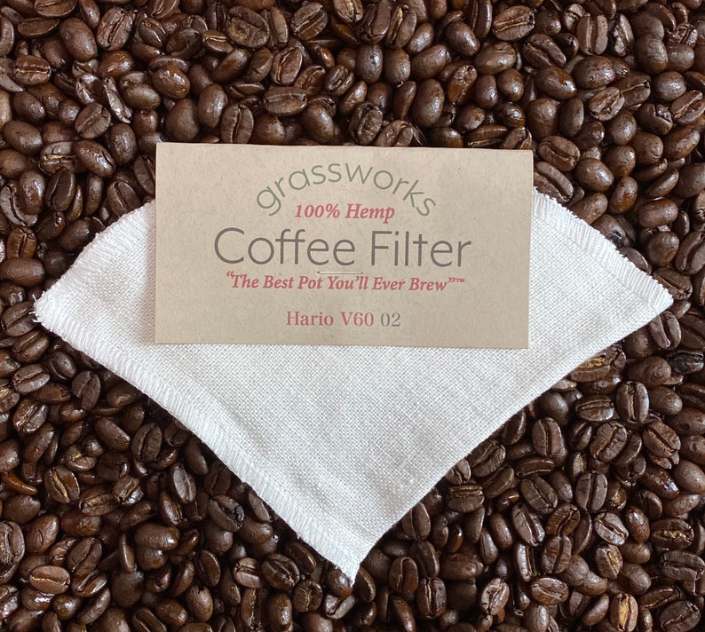 May include: A white hemp coffee filter for a Hario V60 02 coffee maker. The filter is on a bed of coffee beans. The text on the label reads "grassworks 100% Hemp Coffee Filter "The Best Pot You'll Ever Brew" Hario V60 02"