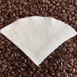 May include: White reusable coffee filter made of fabric, lying on a bed of brown coffee beans.