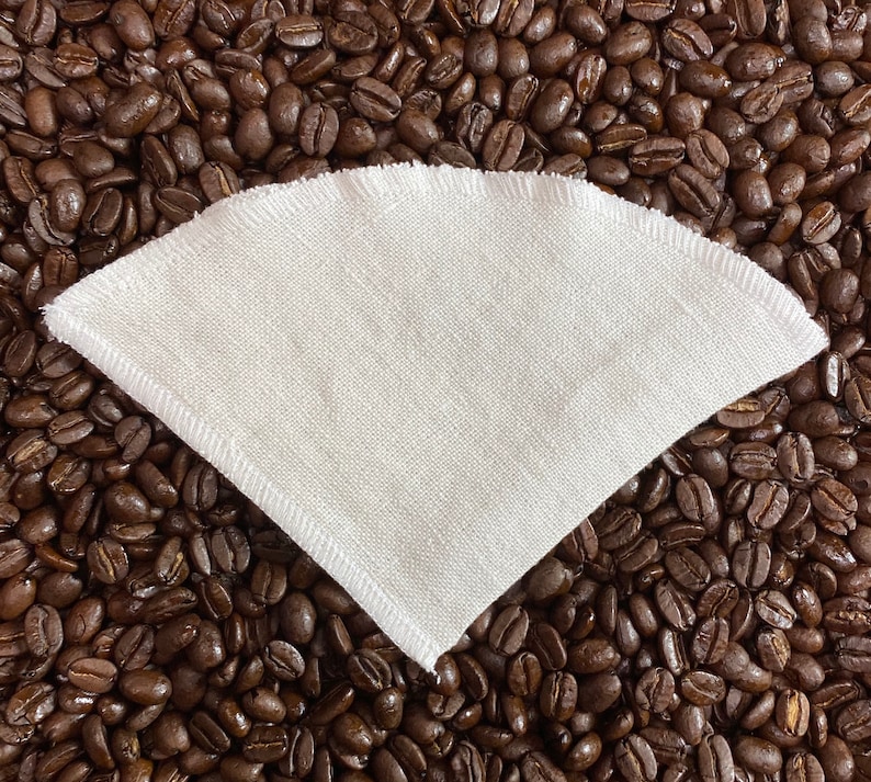May include: White reusable coffee filter made of fabric, lying on a bed of brown coffee beans.