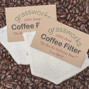 Puede incluir: Dos filtros de café cónicos de color blanquecino con el texto "grassworks 100% Hemp Coffee Filter" en una tarjeta marrón. Los filtros están sobre un lecho de granos de café. El texto de la tarjeta dice "The Best Pot You'll Ever Brew".