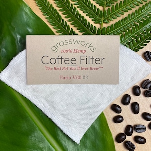 May include: A white hemp coffee filter for a Hario V60 02 coffee maker. The filter is on a wooden surface with coffee beans scattered around it. The text on the filter reads "grassworks 100% Hemp Coffee Filter "The Best Pot You'll Ever Brew" Hario V60 02".