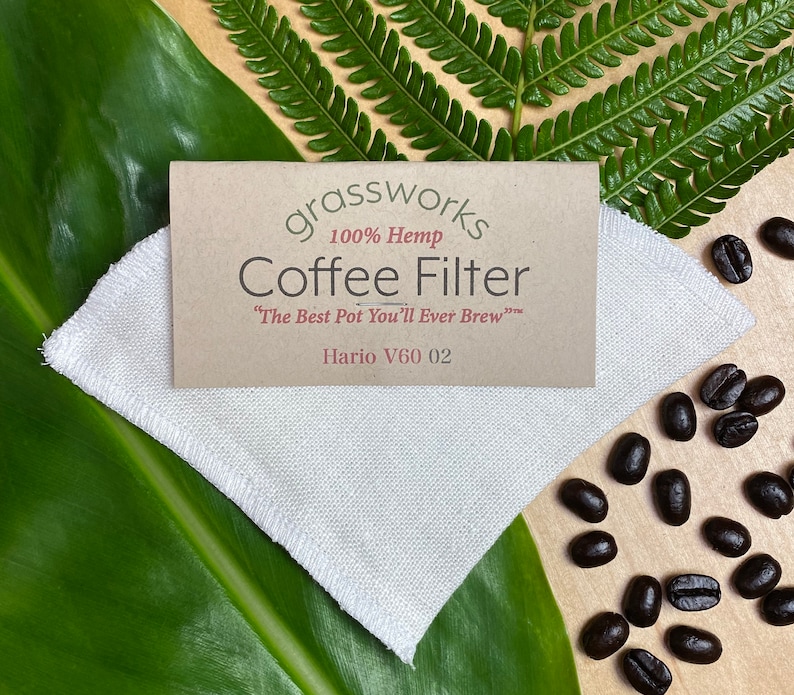 May include: A white hemp coffee filter for a Hario V60 02 coffee maker. The filter is on a wooden surface with coffee beans scattered around it. The text on the filter reads "grassworks 100% Hemp Coffee Filter "The Best Pot You'll Ever Brew" Hario V60 02".