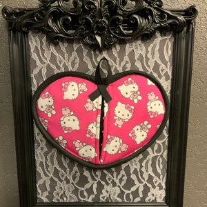 May include: A pink heart-shaped oven mitt with a black trim and a pattern of Hello Kitty characters. The mitt is hanging on a black ornate frame with a white lace background.