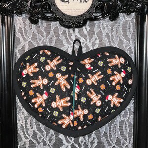 May include: A black heart-shaped oven mitt with a pattern of gingerbread men with skeleton bones, wearing Santa hats and holding candy canes. The background is black with white sprinkles.