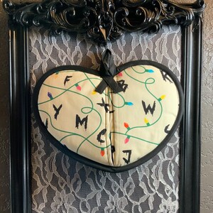 May include: A heart-shaped oven mitt with a white background and a green and black pattern of Christmas lights and letters. The letters spell out "F Y M C W J".