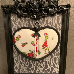 May include: A heart-shaped oven mitt with a black trim and a white background featuring a Grinch Christmas design. The mitt is displayed in a black frame with a white lace background.