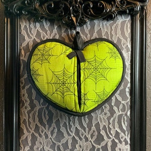 May include: A green heart-shaped pillow with a black spiderweb pattern hanging in a black ornate frame with white lace behind it.