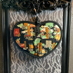 May include: A heart-shaped black and green pot holder with a tiki-themed design. The pot holder features images of palm trees, tiki gods, and women in grass skirts. The text "Exotic Luau" is printed on the pot holder.