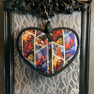 May include: A black framed heart-shaped pot holder with a Spiderman comic book design. The pot holder is hanging on a black lace background.