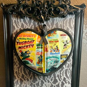 May include: A black framed heart-shaped picture with a white lace background. The heart is made of fabric with a collage of vintage Mickey Mouse comic book covers. The text on the covers includes "ALT DISNEY'S Mickey Mouse", "TUGBOAT MICKEY", "MICKEY SHINES", "WALT DISNEY'S MICKEY MOUSE", "WALT DISNEY", and "MICKEY MOUSE".
