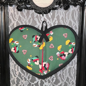 May include: Green heart-shaped pot holder with a black trim. The pot holder features a repeating pattern of Mickey Mouse dressed as an elf kissing Minnie Mouse. The pot holder has a black ribbon loop for hanging.