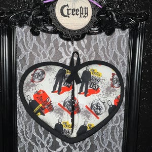 May include: A black framed heart-shaped pot holder with a white background and a black and white print of a horror movie character. The print includes the text "The Back", "The Night", "He's Back", "Stay Alive", and "One Last Time".