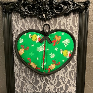 May include: Green heart-shaped oven mitt with a black trim. The mitt features a Christmas-themed print with the Grinch and Cindy Lou Who.