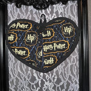 May include: A black heart-shaped pot holder with a Harry Potter theme. The pot holder is decorated with a black and gold diamond pattern and the words "Harry Potter" and "HP" in gold lettering.