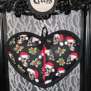 May include: A black heart-shaped pot holder with a pattern of skulls wearing Santa hats and holly leaves. The pot holder has a red ribbon attached to the top.
