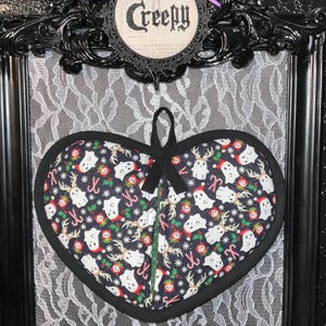 May include: A black heart-shaped pot holder with a festive pattern of ghosts wearing Santa hats, candy canes, and reindeer antlers. The pot holder is hanging in a black frame with white lace behind it. The text "Creepy" is written on a circular tag above the pot holder.