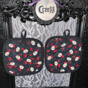 May include: A pair of black quilted pot holders with a red and white pattern of a horror movie character's mask. The pot holders have a loop for hanging.