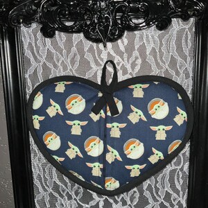 May include: Heart-shaped navy blue pot holder featuring a repeating pattern of a cartoon character. The pot holder has a black trim and a black loop for hanging. A black bow is tied at the top. The pot holder is displayed against a black framed background.