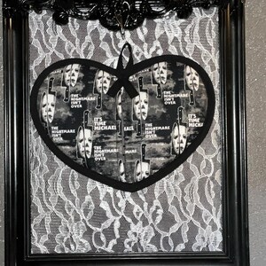 May include: Black framed wall art featuring a heart-shaped design with a horror movie theme. The heart-shaped design has a repeating pattern of a mask and knife with the text "The Nightmare Isn't Over" and "It's Time Michael". The background is white lace.