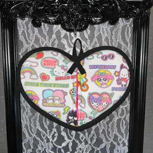 May include: A heart-shaped, quilted pot holder featuring Hello Kitty and other Sanrio characters. The pot holder has a black border and a black ribbon for hanging. It is set against a black framed background with white lace.