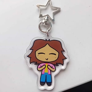 Peut inclure: Un porte-clés en acrylique transparent représentant un personnage de dessin animé avec des cheveux bruns, une chemise rose, un sous-vêtement jaune, un jean bleu et une expression joyeuse. Le porte-clés est attaché à un anneau en forme d'étoile argenté.