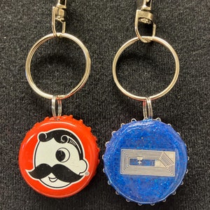 May include: Two keychains with bottle cap charms. One charm is red with a cartoon man with a mustache. The other charm is blue with a silver glitter design.