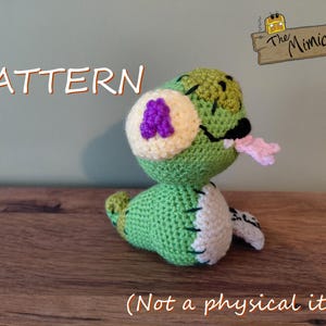 Op de afbeelding: Een groen gehaakt wezen met een crèmekleurige buik, een roze tong en een paarse vlek op de zijkant. De tekst .pdf PATTERN (Not a physical item) en The Mimicry zijn zichtbaar. Het wezen heeft zwarte gestikte details en een textuurrijk oppervlak.