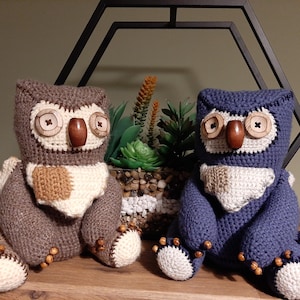 May include: Two crocheted owl plushies, one brown and one blue, with wooden button eyes. The owls are sitting on a wooden surface.
