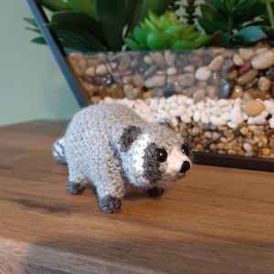 May include: A crocheted raccoon figure in grey, black and white. The small animal has a striped tail and is positioned on a wooden surface. The background includes a decorative planter with stones and greenery.