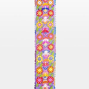 May include: A long, rectangular, embroidered textile with a lavender background. The design features colorful flowers in shades of red, yellow, orange, and pink, with green leaves. The edges are trimmed with a fringe of small, colorful pom-poms.