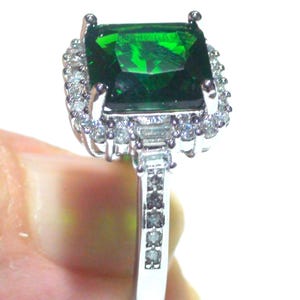 May include: A silver ring with a large, emerald-cut, vivid green gemstone. The gemstone is encircled by a halo of smaller, clear stones. The ring's band is also decorated with smaller, clear stones, creating a sparkling effect.