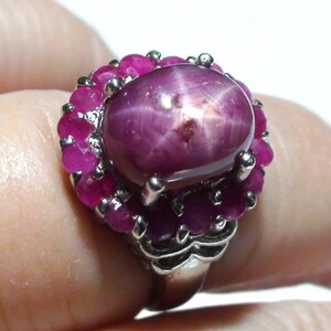 May include: A silver ring featuring a large, oval-shaped, pink star ruby surrounded by smaller pink rubies.
