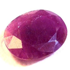 May include: A faceted, oval-shaped ruby gemstone. The gem is a deep magenta colour with a polished surface. The facets catch the light, creating highlights and shadows. The gemstone is set against a white background.
