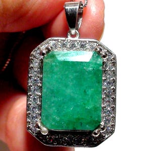 May include: A rectangular emerald pendant with a silver setting and a halo of small, clear gemstones. The emerald is a vibrant green color and has a faceted surface. The pendant hangs from a silver chain.