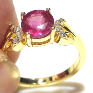 Puede incluir: Un anillo de oro con una gran gema de rubí redonda y facetada. El rubí es de color rojo rosado intenso. El anillo tiene una banda de oro con pequeños diamantes de acento a cada lado del rubí.