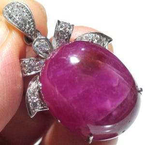 May include: A pendant featuring a large, oval-shaped, deep magenta gemstone. The gemstone is set in a silver-coloured metal with a bow-shaped design, embellished with small, clear gemstones. The pendant is held by a hand.