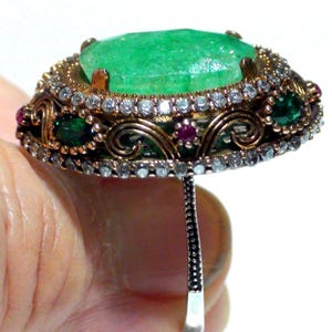 May include: An ornate ring featuring a large, emerald-cut green gemstone as the centerpiece. The ring is surrounded by smaller clear and green stones, with accents of red. The setting has a gold and silver tone, showcasing intricate filigree details.