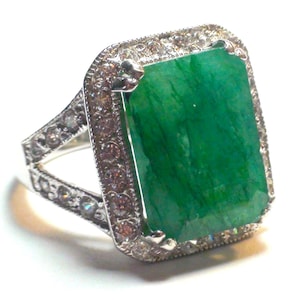 May include: A silver ring featuring a large, rectangular emerald-cut green gemstone. The emerald is surrounded by a halo of small, clear gemstones. The ring's band is also adorned with smaller gemstones, creating a sparkling effect.