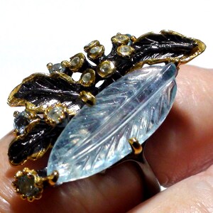 May include: A ring featuring a large, carved, light blue, leaf-shaped gemstone set in gold prongs. The band and decorative elements include dark, oxidized metal leaves accented with small, round, clear stones set in gold-toned metal settings.
