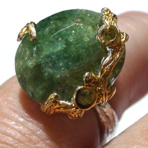 May include: A ring featuring a large, oval-shaped, translucent green gemstone with a textured, organic-looking gold-tone metal setting that wraps around the stone, accented with two small, round, light green stones embedded in the metal branches.