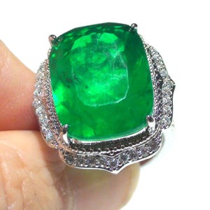 May include: A striking emerald ring featuring a large, square-cut green gemstone. The ring is set in a silver-toned metal and encircled by smaller, clear gemstones. The ring is held up by a hand.