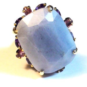 May include: A ring featuring a large, faceted, light blue rectangular gemstone as the centerpiece. The central stone is surrounded by a halo of smaller, oval-shaped, deep purple amethyst gemstones set in gold prongs against a white background.