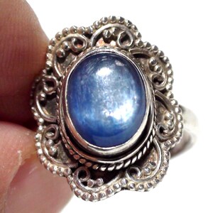 Op de afbeelding: Een zilveren ring met een centrale ovaalvormige blauwe edelsteen. De zetting is omgeven door een decoratieve metalen rand met een geschulpte, kralen afwerking en gedetailleerde draadkrulmotieven die de gepolijste blauwe steen omlijsten.