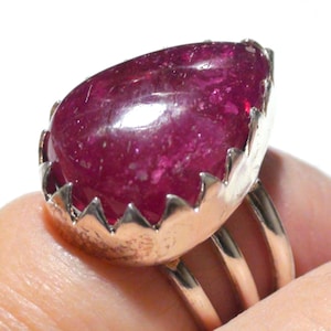 Op de afbeelding: Een zilveren ring met een grote, druppelvormige robijn die in een gegolfde zetting is gezet. De ring heeft drie banden.