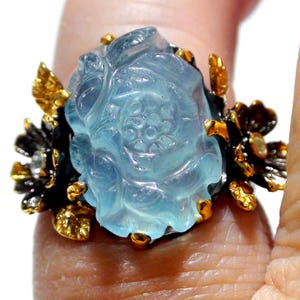 May include: A ring featuring a large, translucent, light blue carved gemstone shaped like a flower. The setting is dark metal with gold-toned accents, including small sculpted leaves and flowers on either side of the central blue stone.