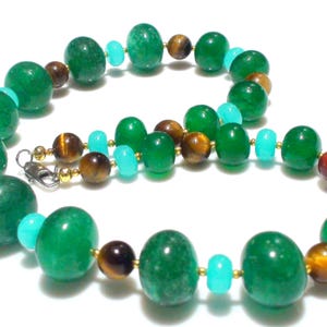 Op de afbeelding: Een kralenketting met grote, ronde, doorschijnende groene kralen afgewisseld met kleinere, gladde, helder turquoise blauwe kralen en bruine, gestreepte tijgeroogkralen. De kralen zijn geregen op een dunne draad met een zilverkleurige metalen sluiting.