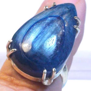 May include: A teardrop-shaped blue kyanite gemstone set in a silver ring. The stone features a polished, smooth surface with visible internal striations and a deep royal blue color. The metal setting includes multiple prongs securing the stone.