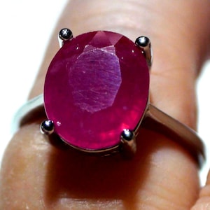 May include: A silver ring featuring a large, oval-cut ruby gemstone. The ruby is a vibrant shade of red with visible facets, held in place by four silver prongs. The ring's band is a simple, polished silver.