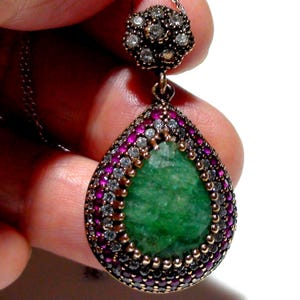 May include: A pendant necklace with a teardrop-shaped design, showcasing a large green gemstone. The gemstone is framed by a border of small red and clear stones. The pendant hangs from a fine silver chain, a sophisticated piece of jewellery.
