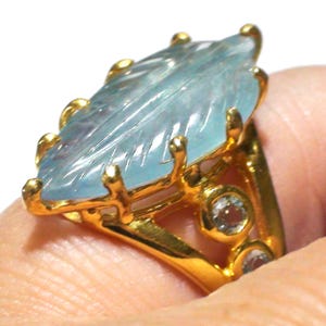 May include: A gold ring featuring a large, marquise-shaped, light blue gemstone carved with a leaf-like pattern. The setting includes multiple prongs securing the central stone, with two smaller, round, light blue accent stones set along the band.